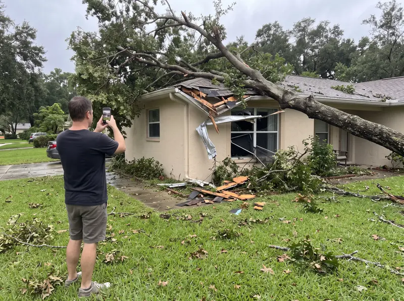 Homeowner on the front lawn photographing a fallen oak that broke through the eave and window of a stucco home